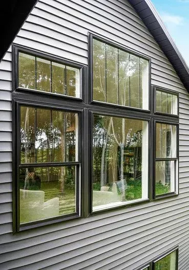 Gray house exterior with black-framed windows reflecting a green yard; sunny day.