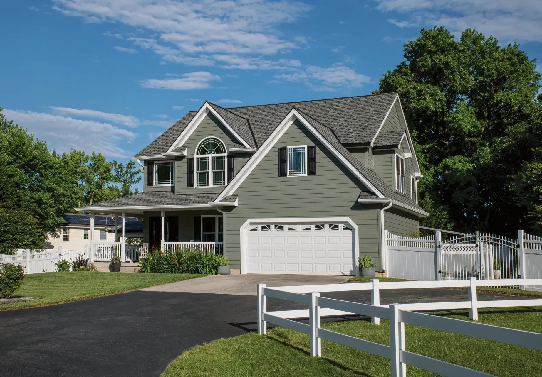 Green two-story house with white garage door, black shutters, and white fence on a sunny day.