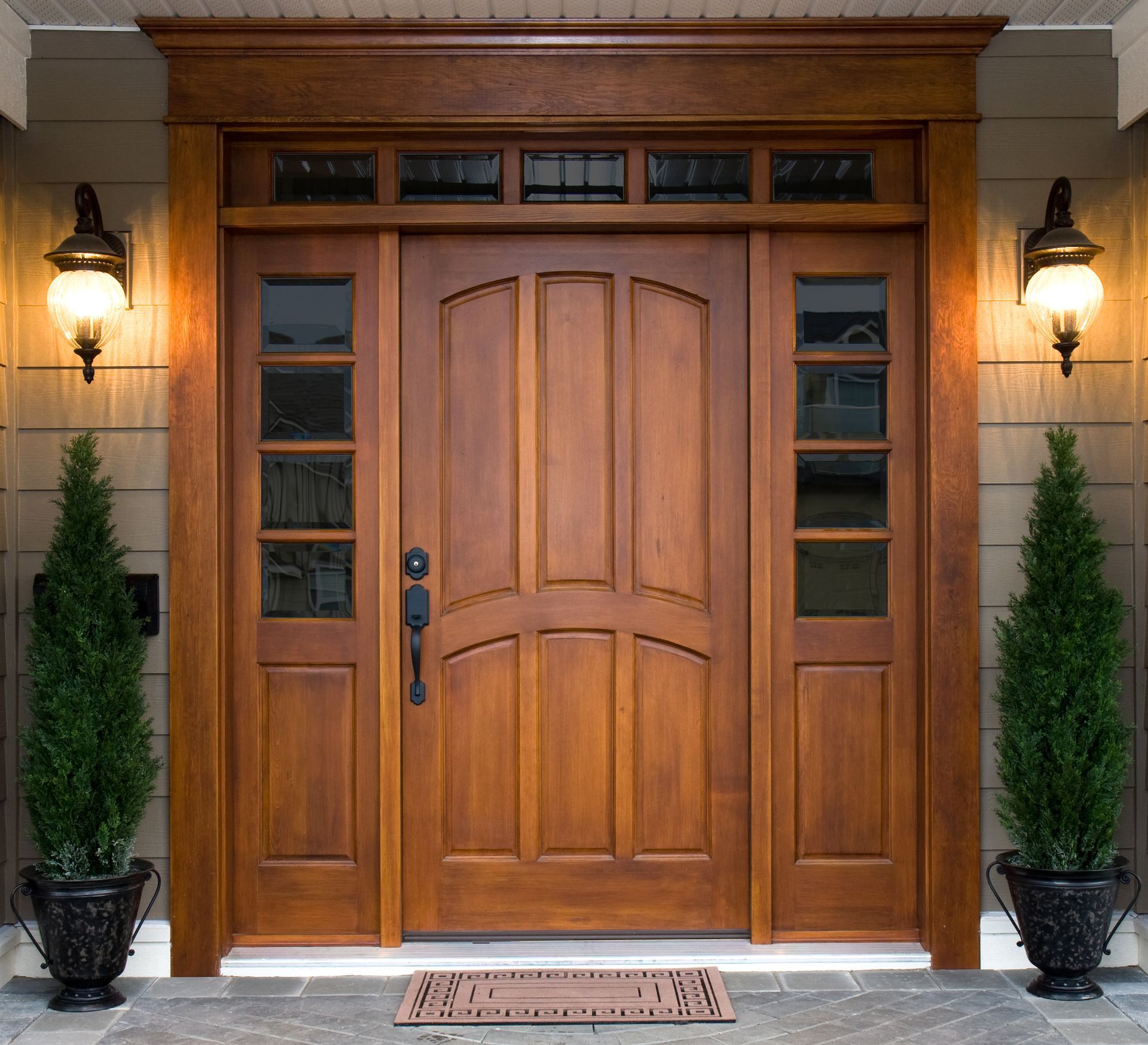 Wooden front door with sidelights, transom windows, sconces, and potted evergreens.