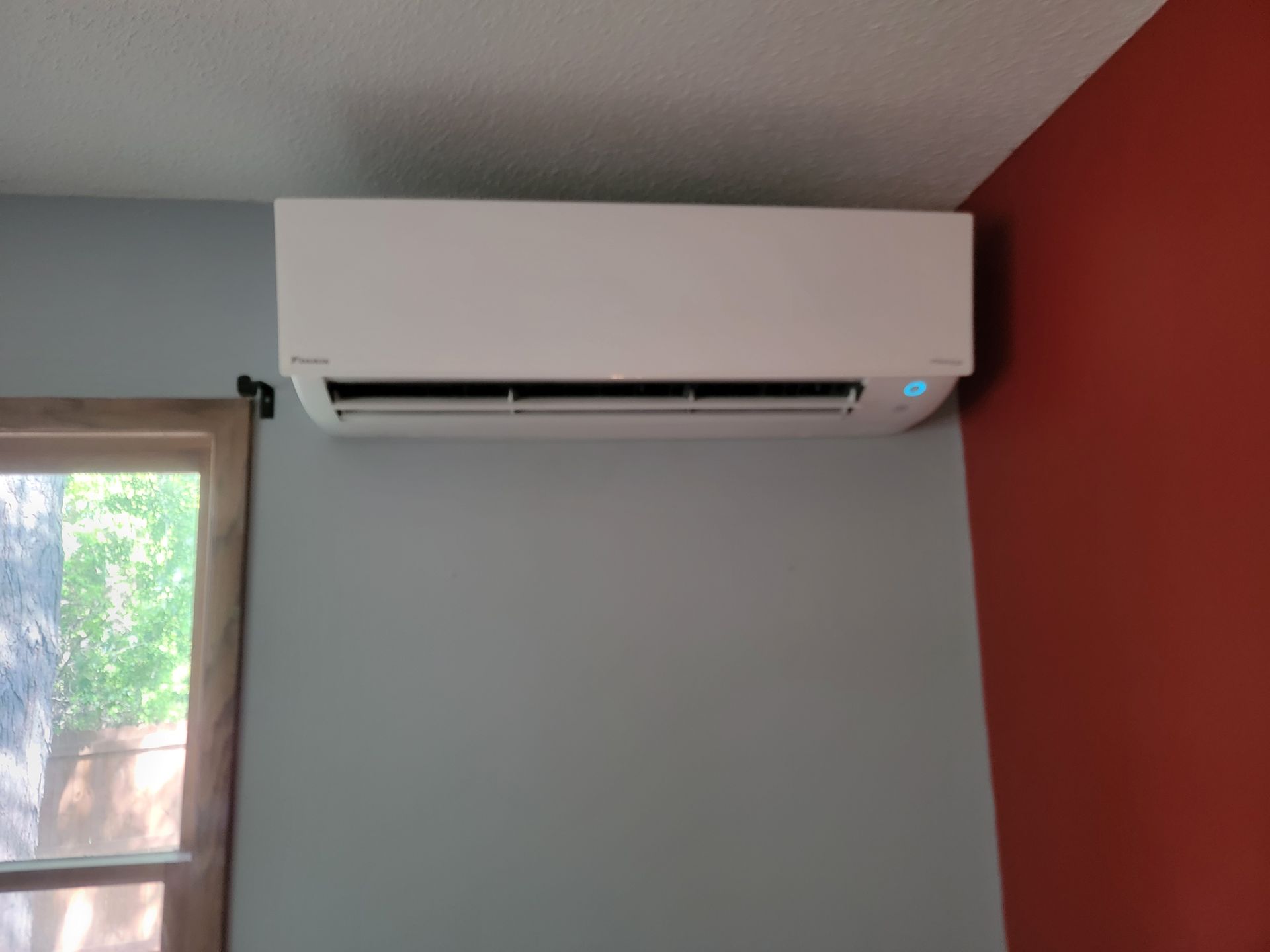 White ductless air conditioner mounted on a gray wall next to a red wall and window.