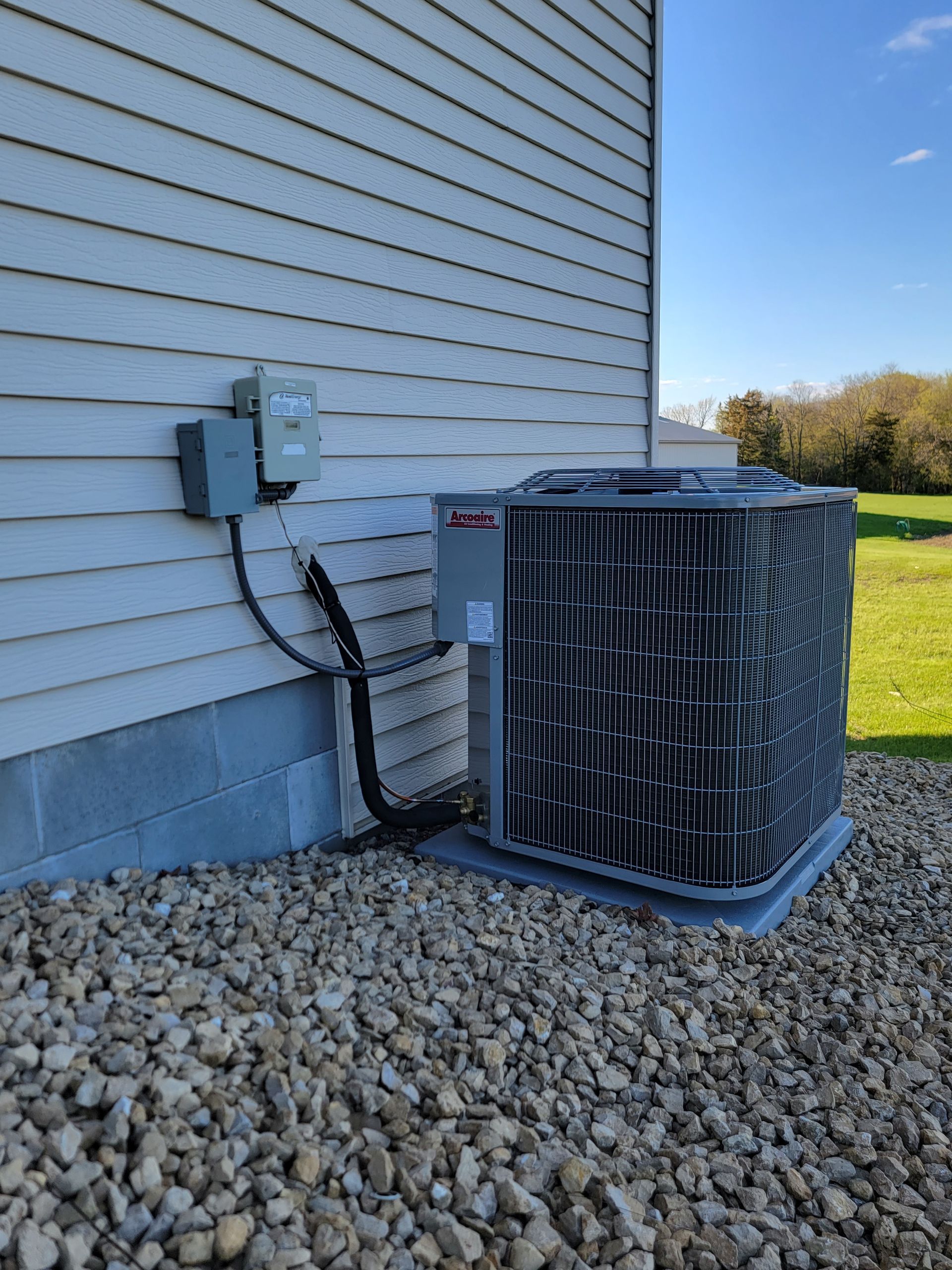 An air conditioning unit outside a beige house, surrounded by gravel, under a blue sky.