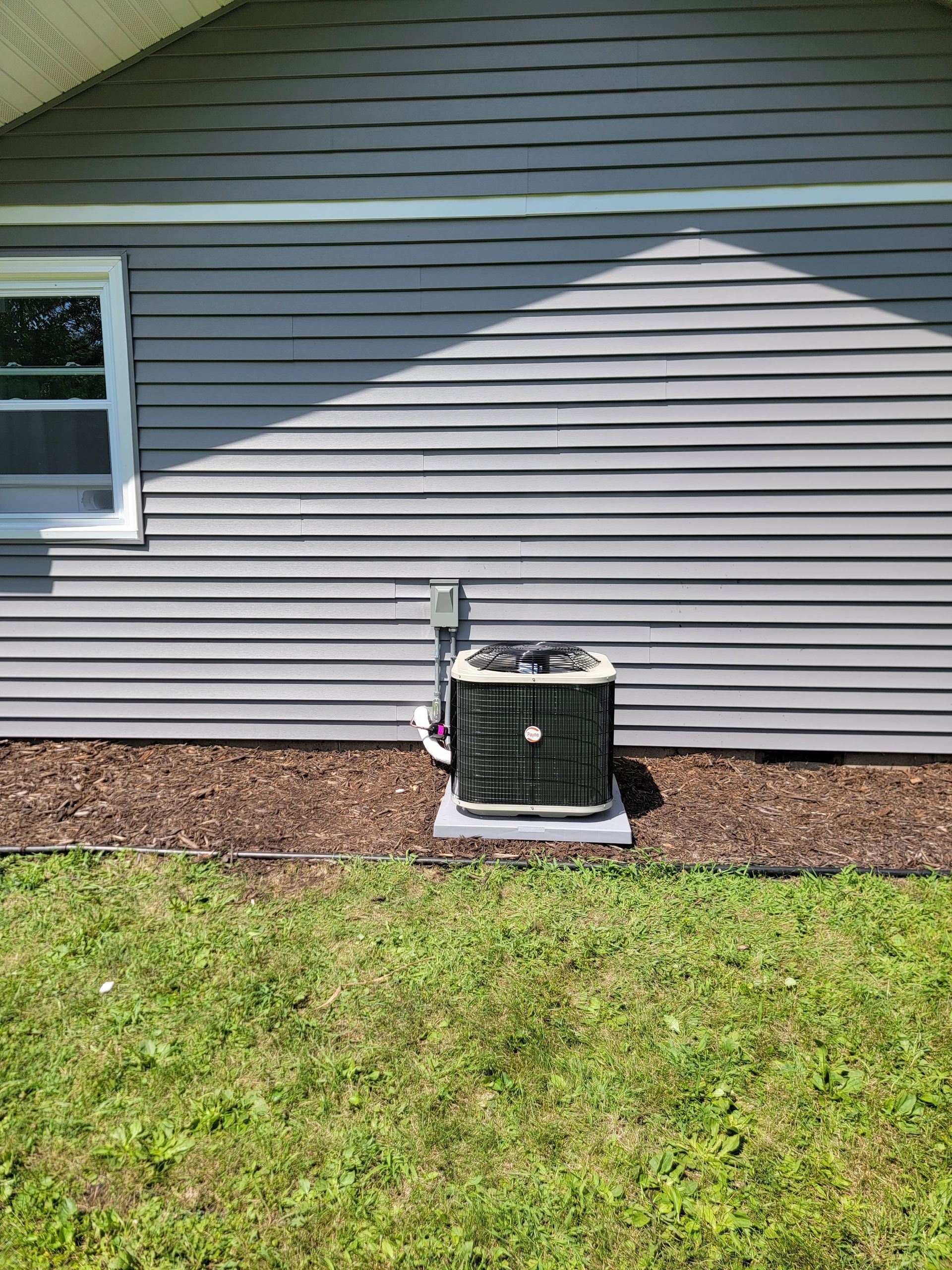 Air conditioner on concrete slab next to gray siding and a window.