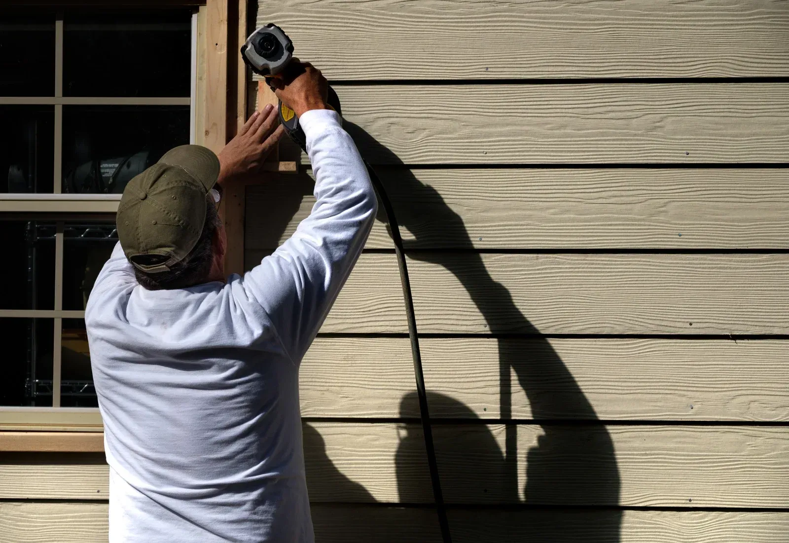 Person drilling into tan siding beside a window, with a cord hanging down.