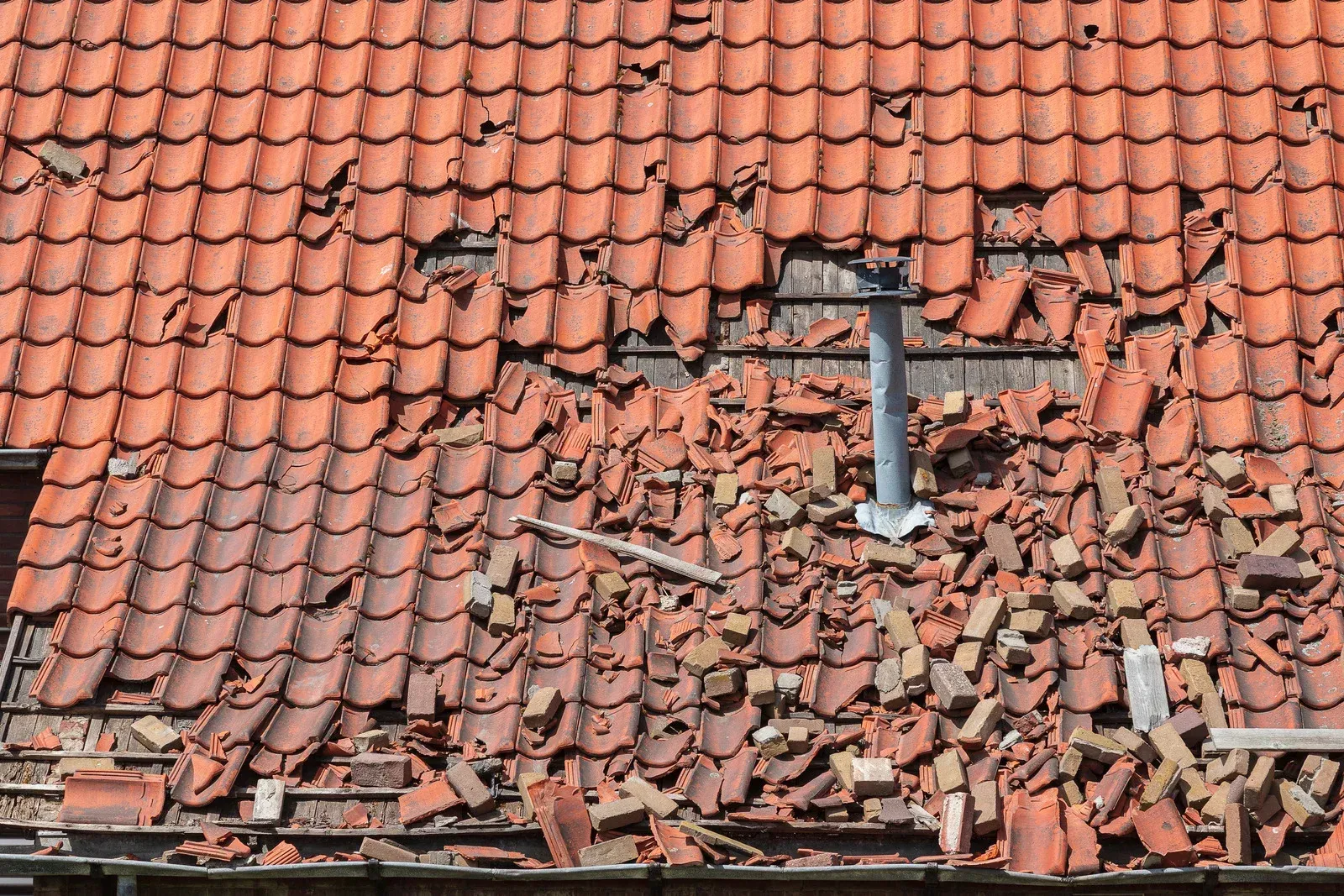 Damaged red-tile roof with a large hole and scattered broken tiles around a chimney vent