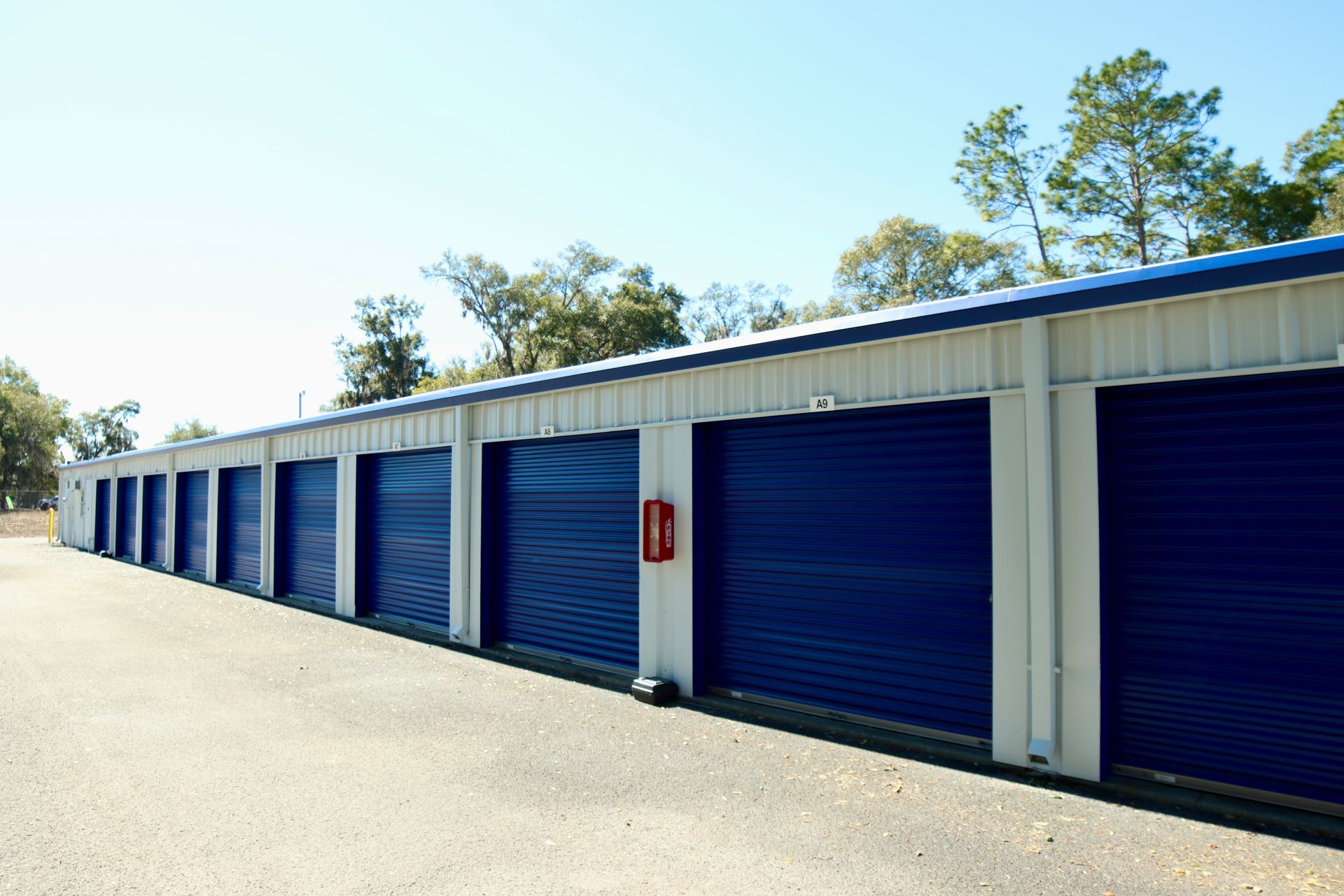 Row of blue storage unit doors under a white and blue metal roof on a sunny day.