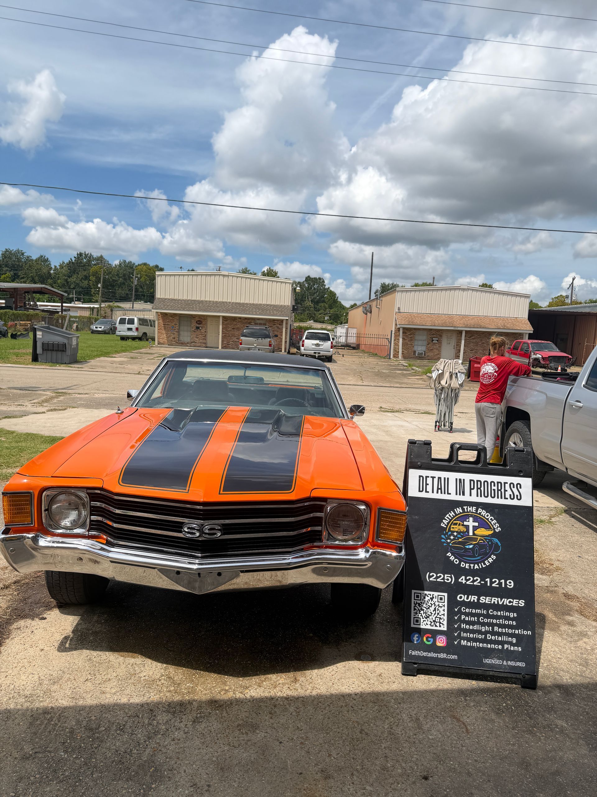Orange classic car with black racing stripes on display at an outdoor car show, with people nearby.