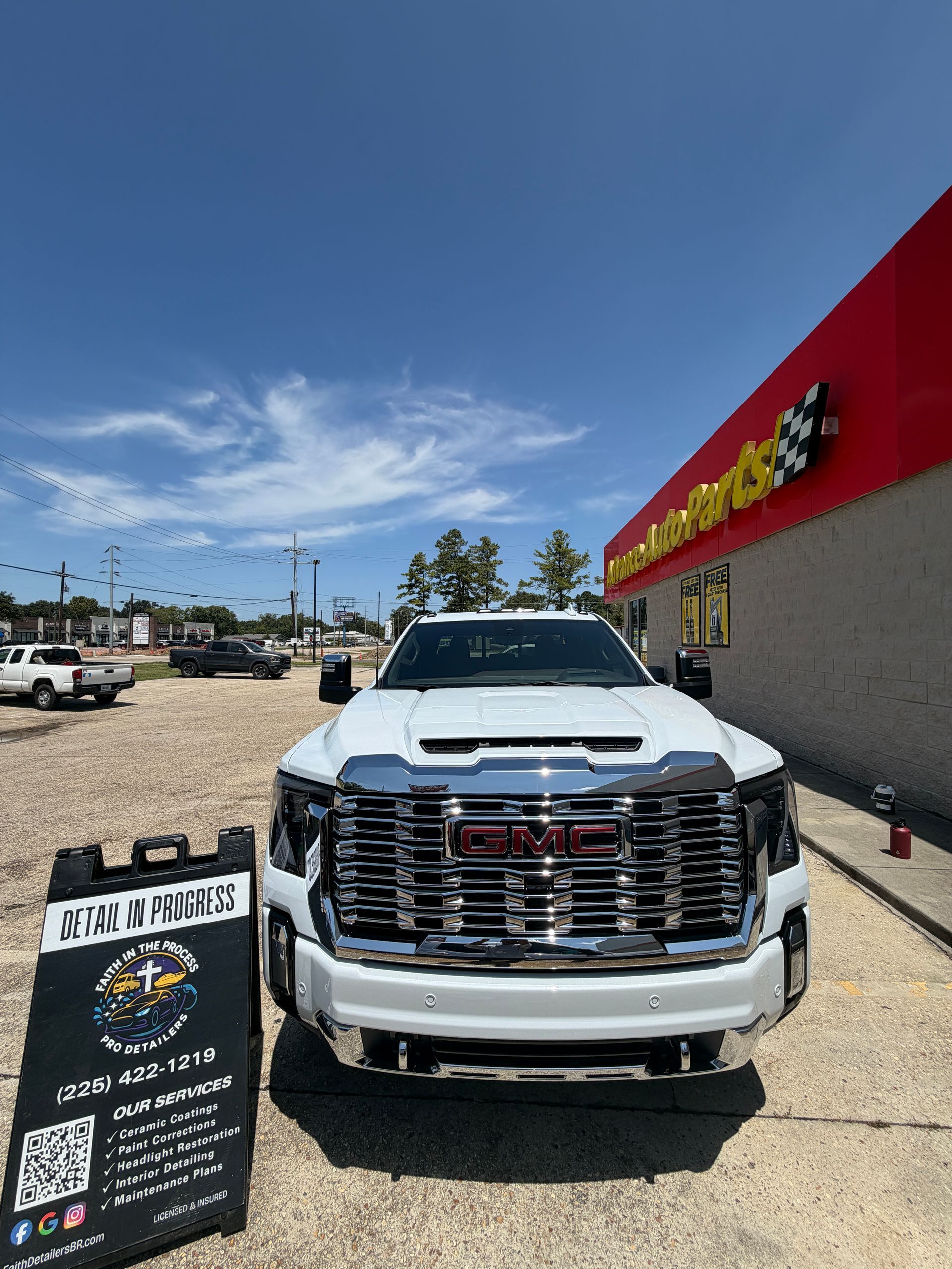 White pickup truck parked outside a car dealership under a clear blue sky, with a promotional sign in front.