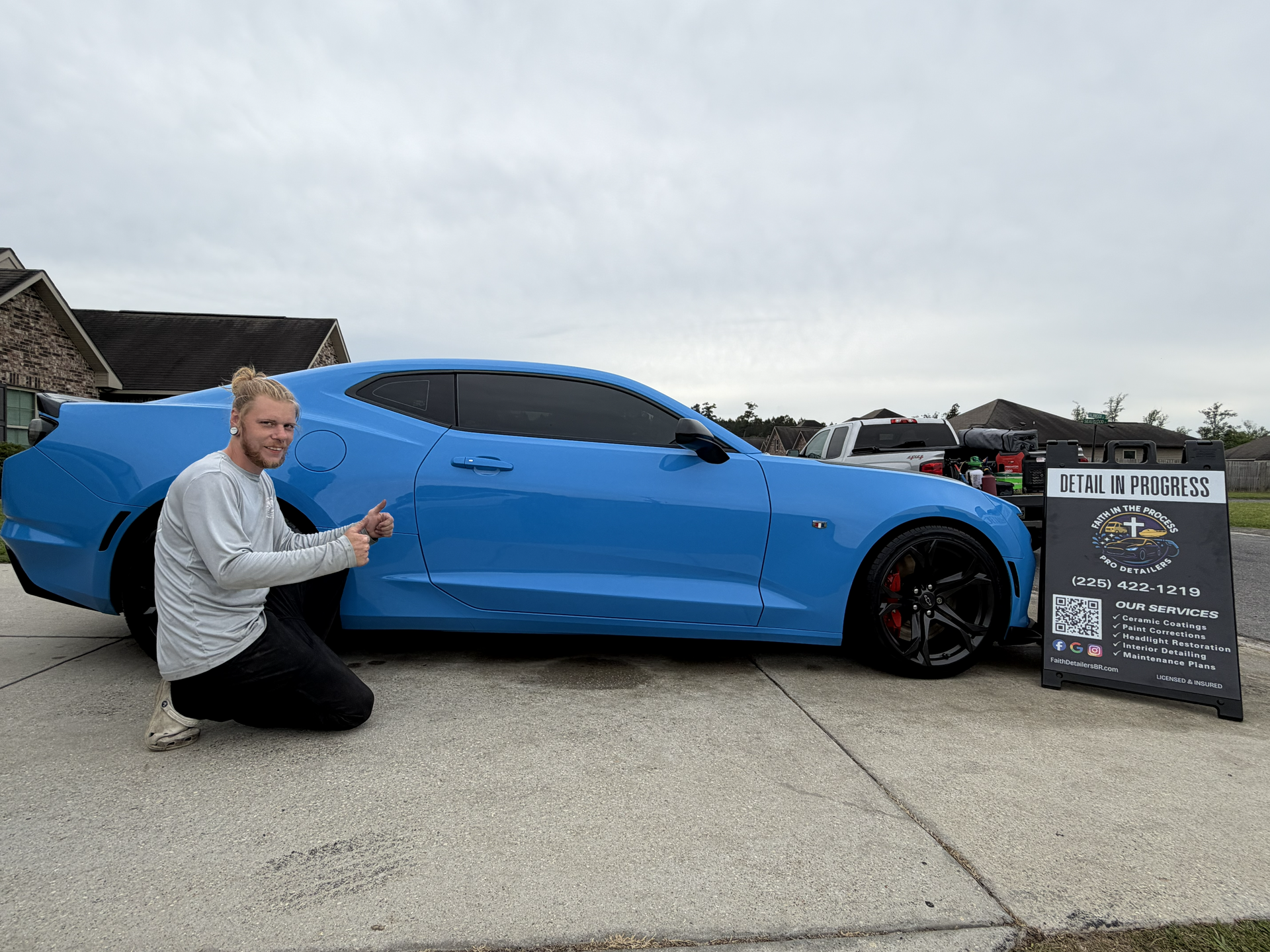 Person kneeling beside a blue sports car with tinted windows and a signboard on a paved lot