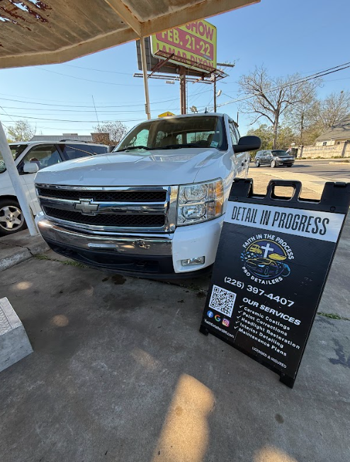 White pickup truck parked by a repair sign reading “Vehicle in Progress” under a canopy.