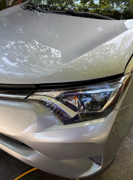 Close-up of a white car’s front headlight and hood in daylight, with tree reflections.