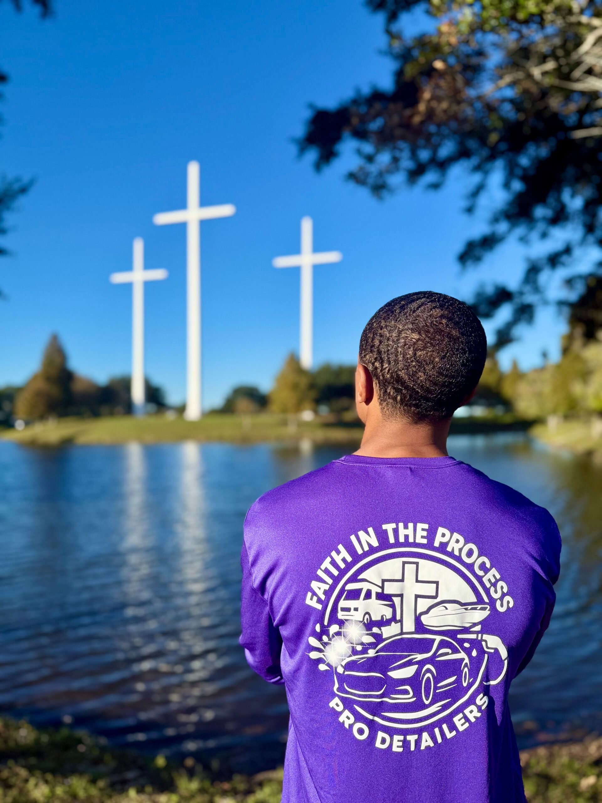 Person in purple shirt facing three white crosses by a lake under a blue sky
