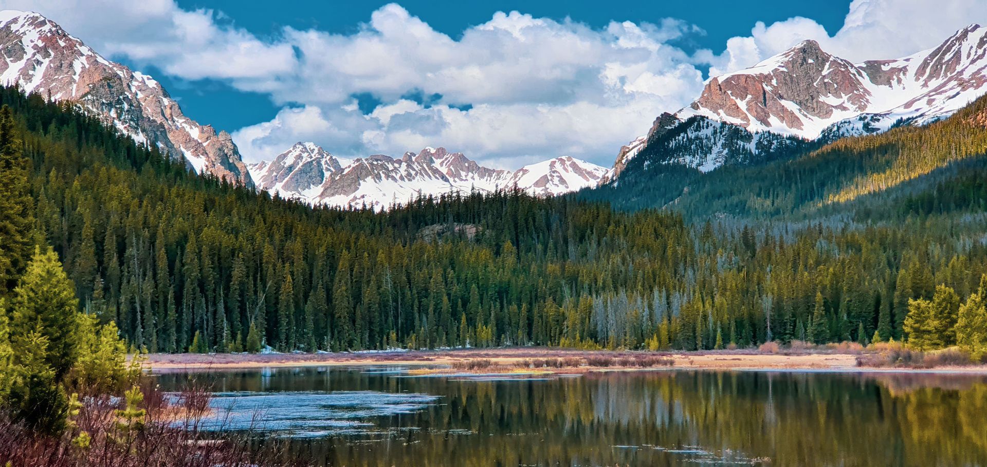 Snowy mountain peaks above a pine forest and calm lake under a blue sky with clouds