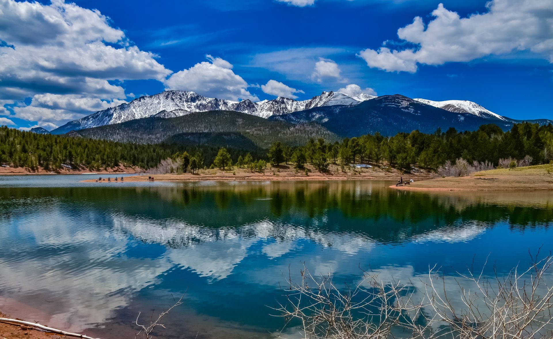Snow-capped mountains under a blue sky, reflected in a calm turquoise lake with forested shoreline.