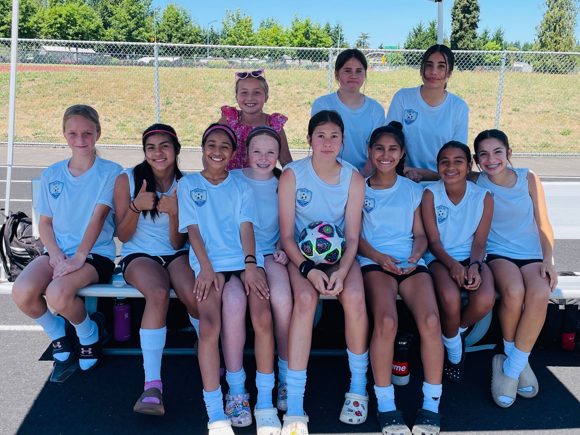 Soccer team of girls in light blue jerseys poses on a bench on a sunny day.