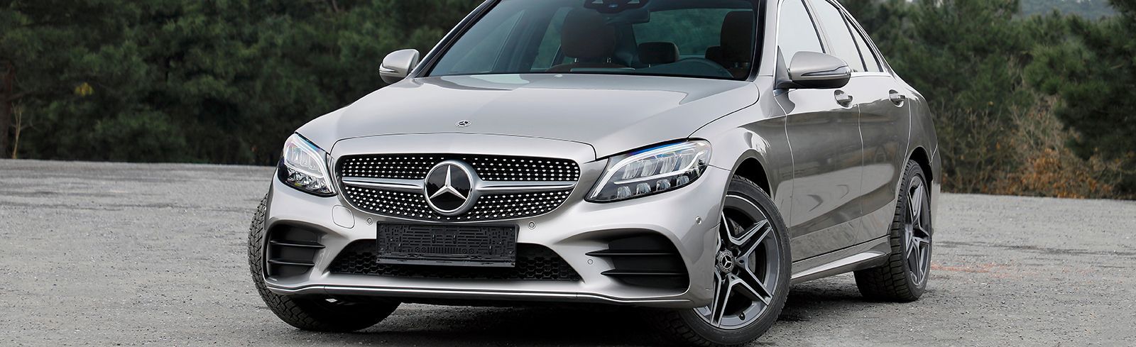 Silver Mercedes-Benz sedan parked on gravel road with blurred trees in background.