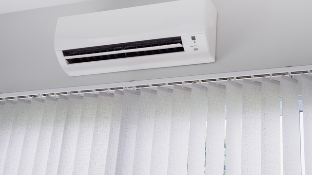 White wall-mounted air conditioner above vertical white blinds on a light-colored wall