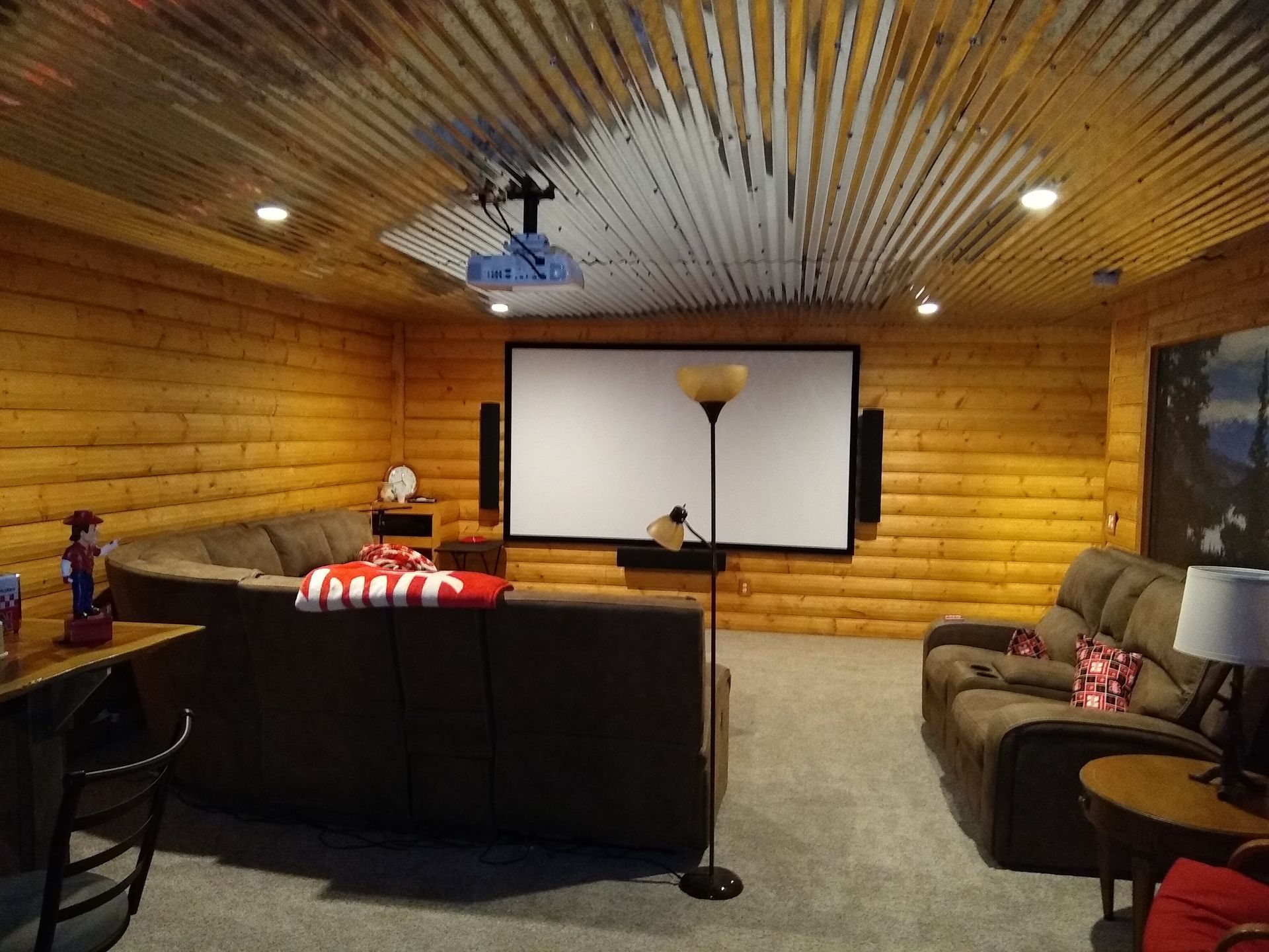 Home theater with a large screen, log walls, and a corrugated metal ceiling. Two couches and a floor lamp are present.
