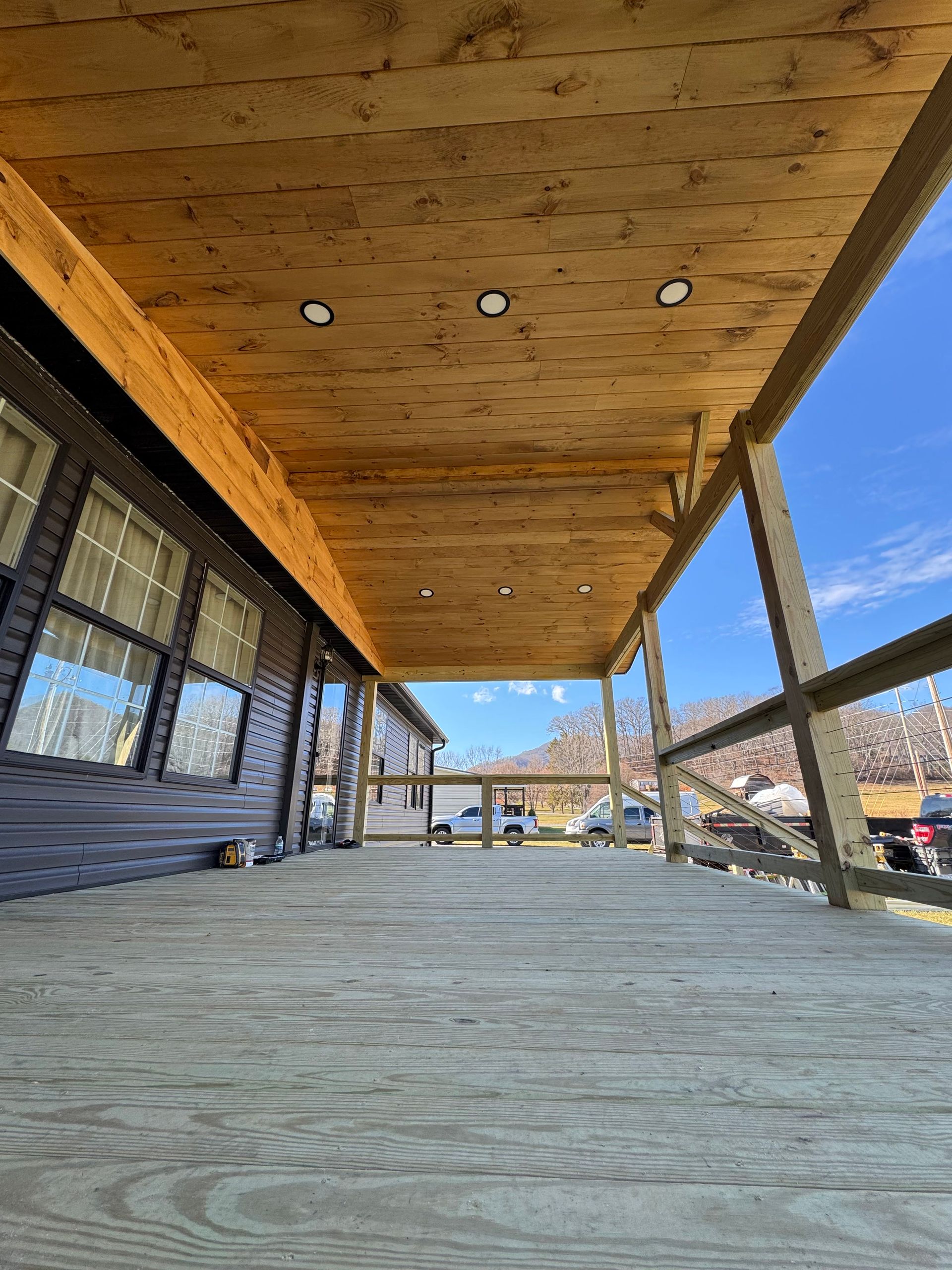 A low-angle view of a wooden porch with recessed lighting, looking out towards a rural landscape under a clear blue sky.