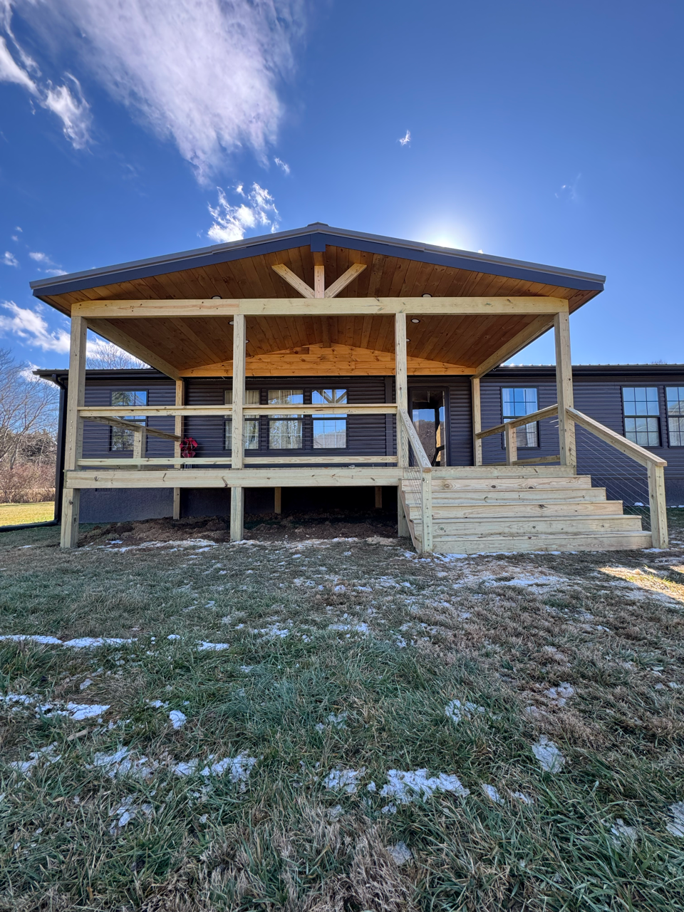A new, wood-framed covered porch addition attached to the side of a dark gray house under a clear, sunny blue sky.