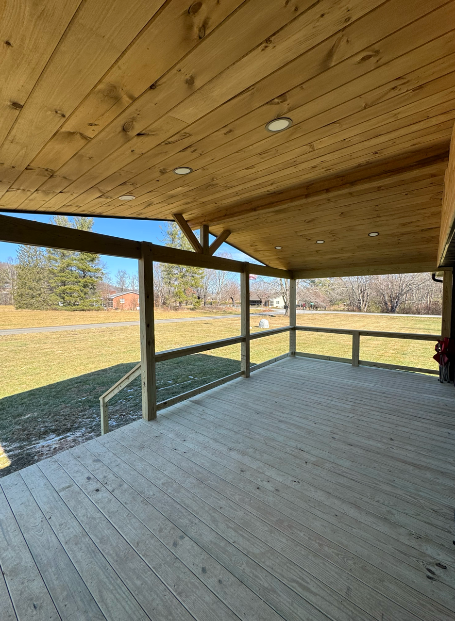 A view from a wooden covered porch looking out onto a grassy lawn and trees under a clear blue sky.