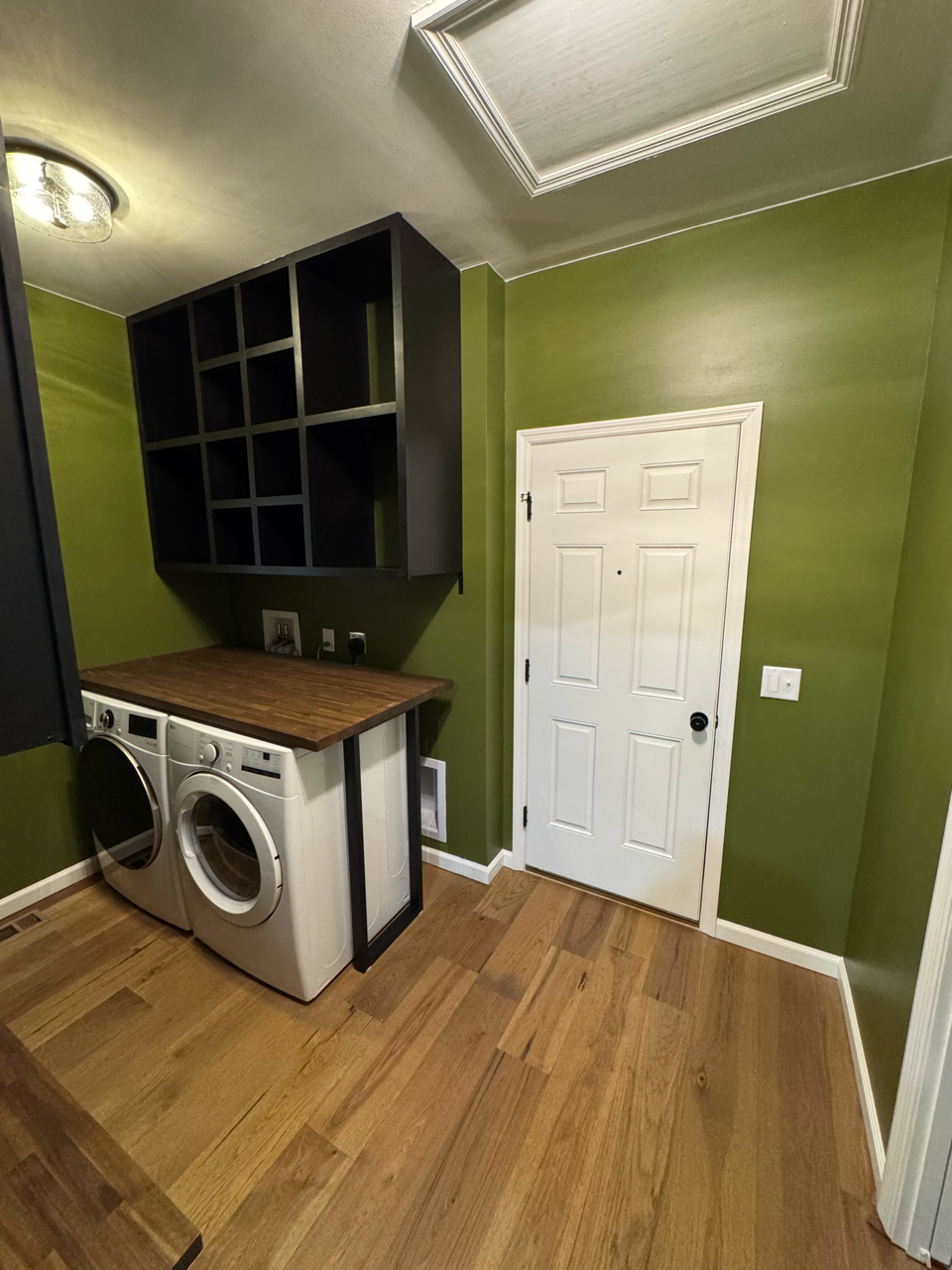 A laundry room with green walls, wood floors, a stacked washer and dryer set with a wooden counter, and black wall cubbies.