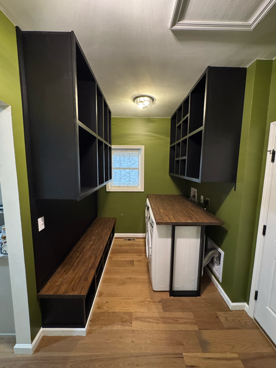 Laundry room with olive green walls, wood floors, black built-in shelving, a wood-topped bench, and a laundry setup.