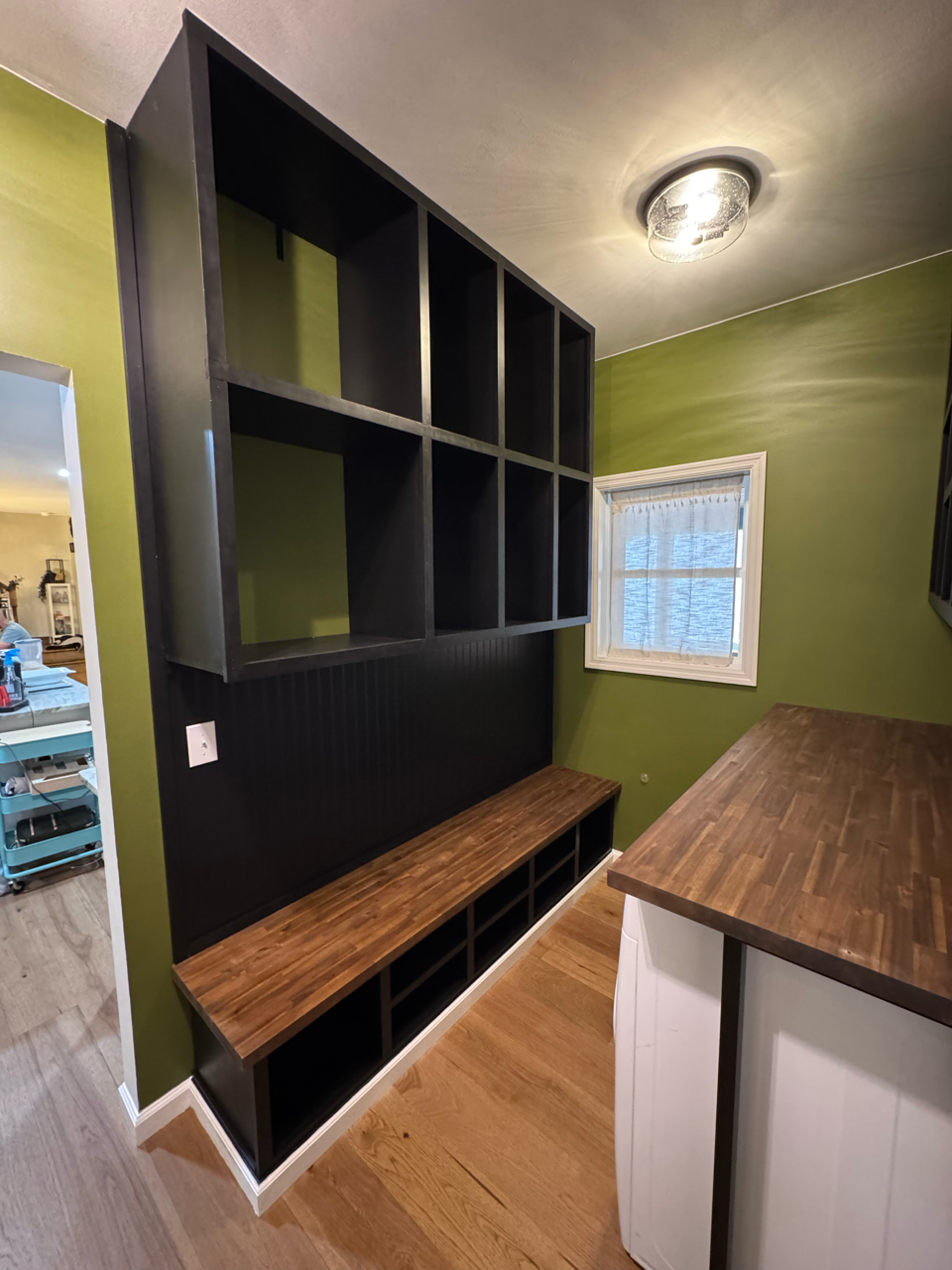 A mudroom entryway featuring black wall-mounted cubbies and a matching bench with a stained wood top against olive walls.