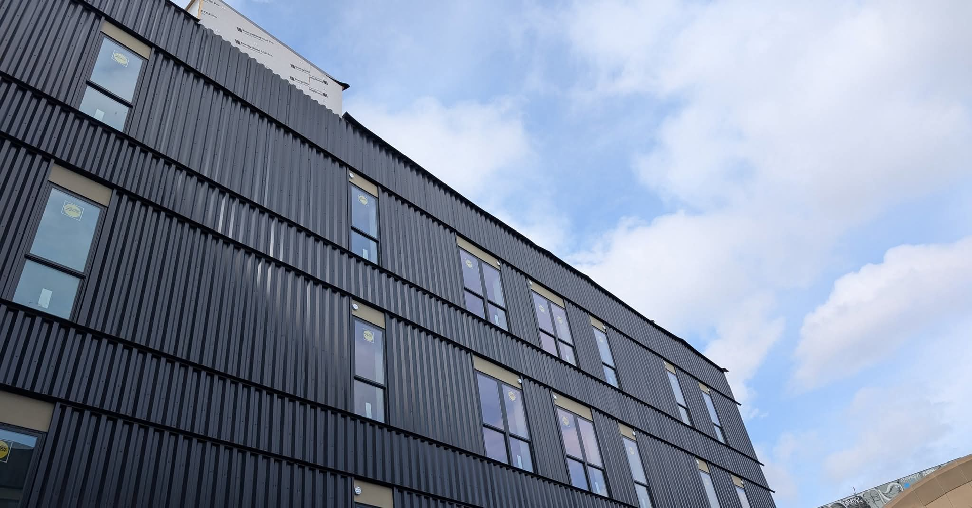 A low-angle view of a modern building with dark, vertical siding and rows of rectangular windows under a cloudy blue sky.