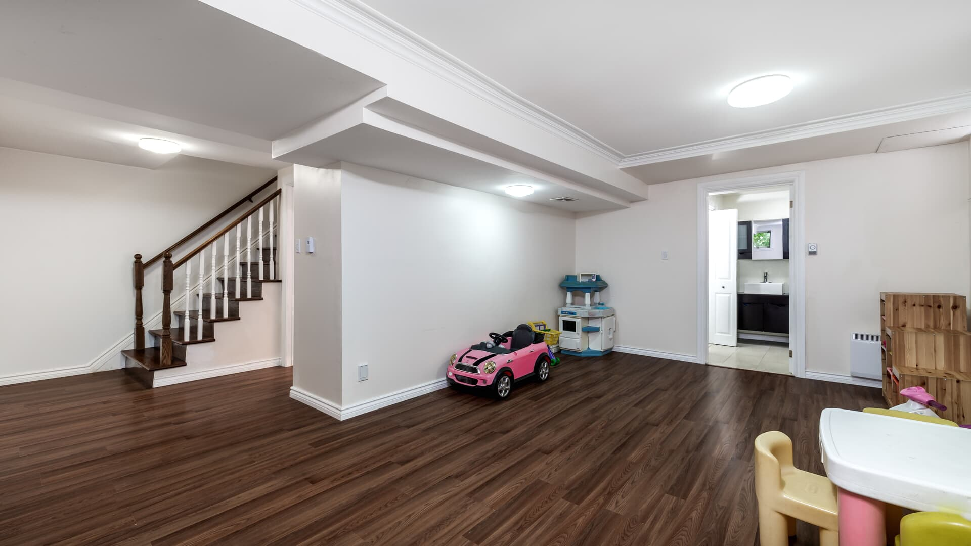A wide, furnished basement room with wood floors, a staircase on the left, a toy car, a play set, and a small table.
