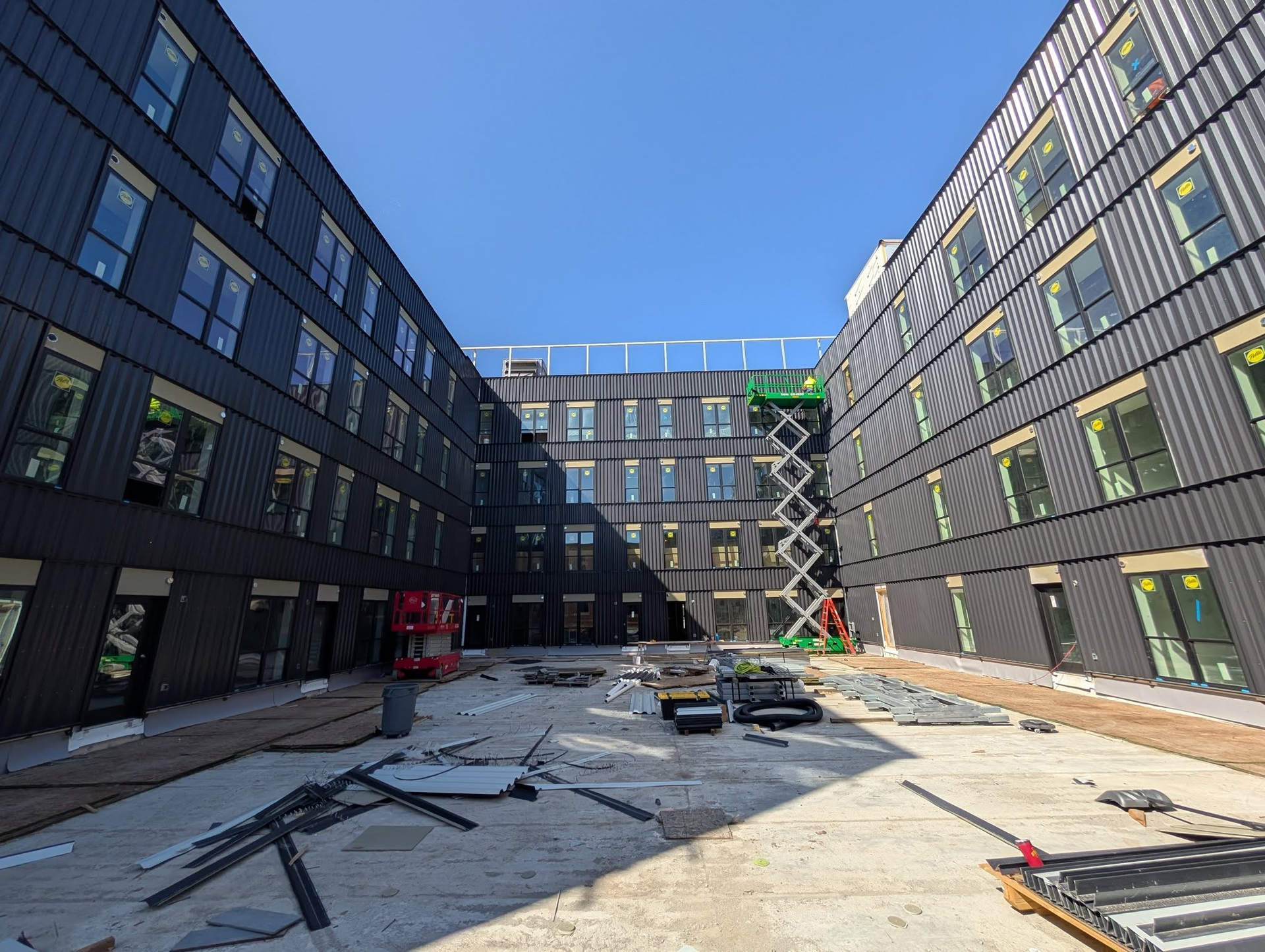 A construction site with two modern, dark-paneled buildings flanking a gravel courtyard and a green scissor lift.