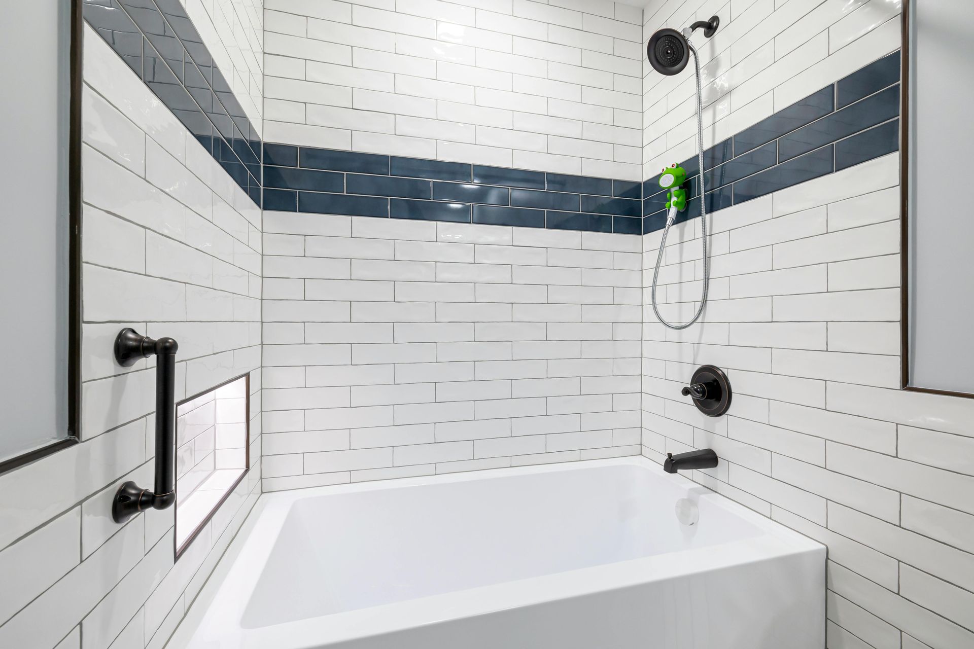 A bathtub with a shower head, a black grab bar, and walls featuring white subway tiles with a horizontal dark blue stripe.