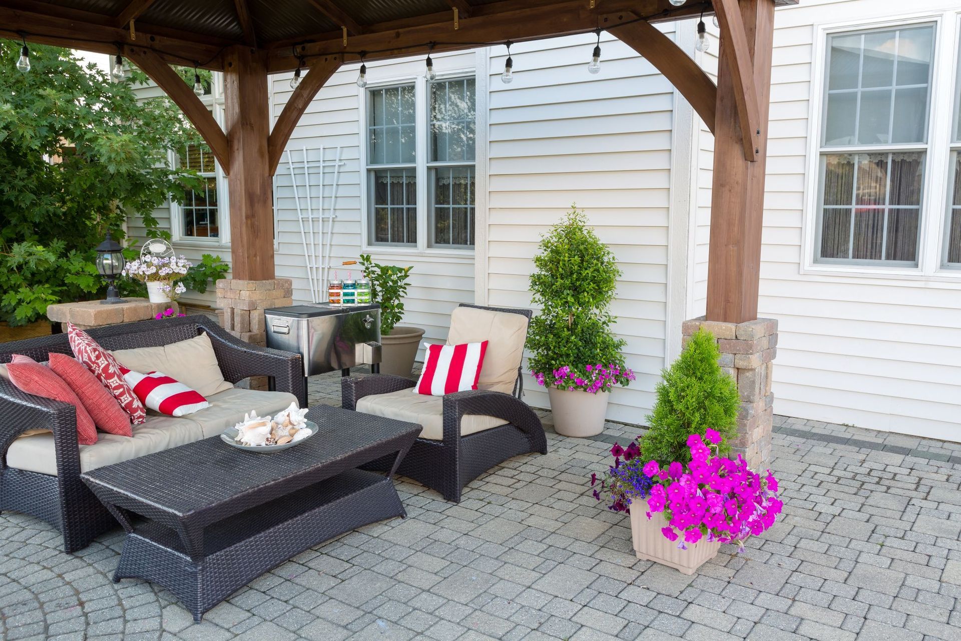 A patio with dark wicker furniture, a wooden pergola, and potted plants with pink flowers against a white house wall.