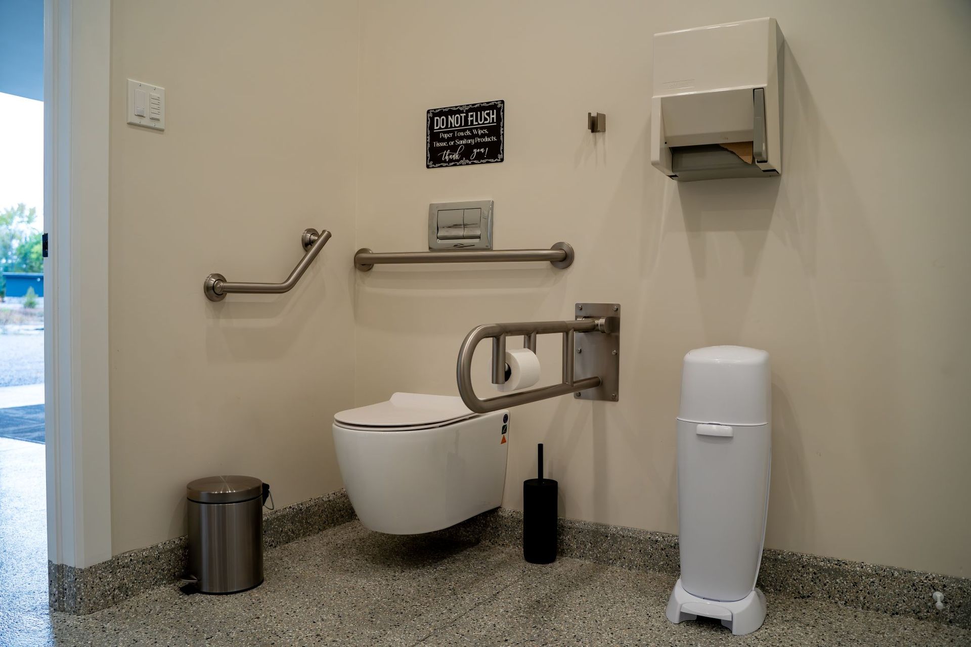An accessible restroom stall with a wall-mounted toilet, grab bars, toilet paper dispenser, and waste bins on a tiled floor.