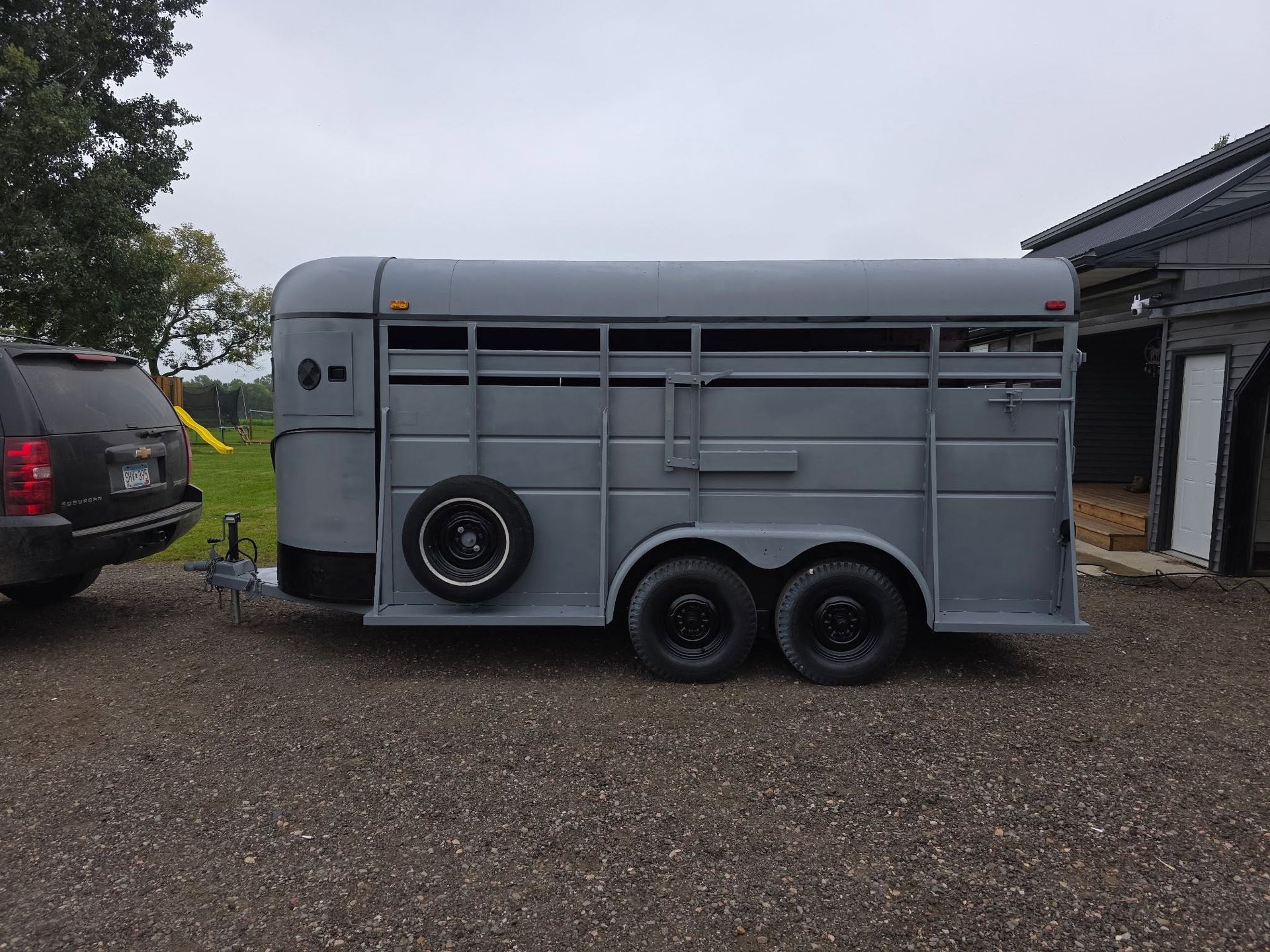 Gray horse trailer parked on gravel beside a building, with a spare tire mounted on the side.