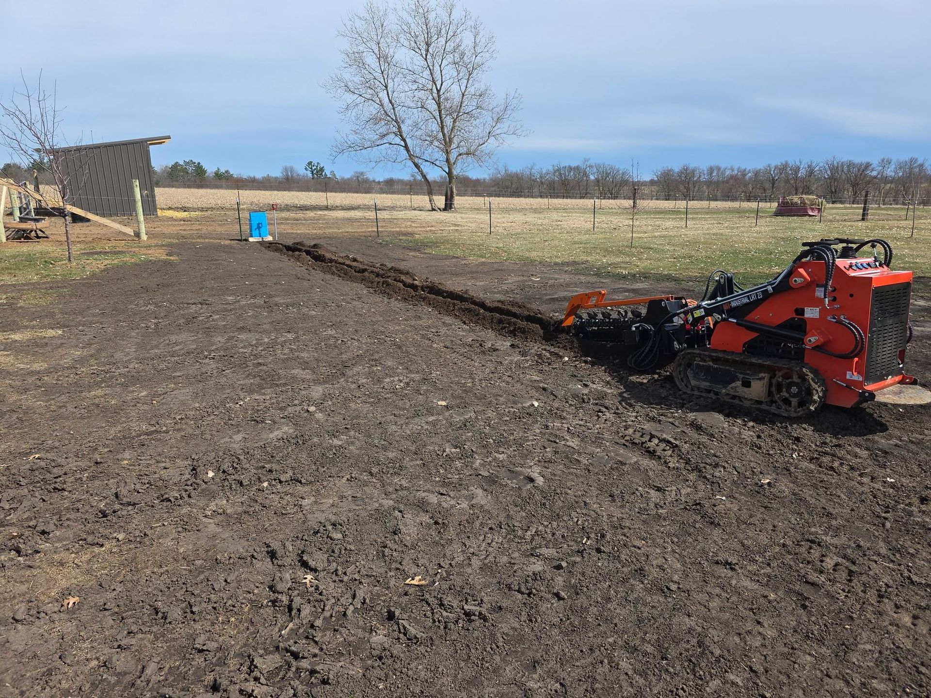 Red tractor tilling a muddy field near a barn under a clear sky