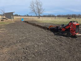 Red tractor tilling a muddy field near a barn under a clear sky