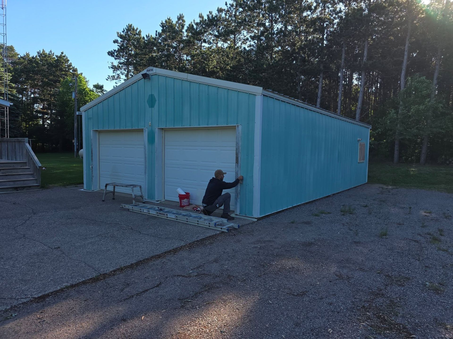 Blue garage with two white doors and a person sitting beside one door on a gravel lot.