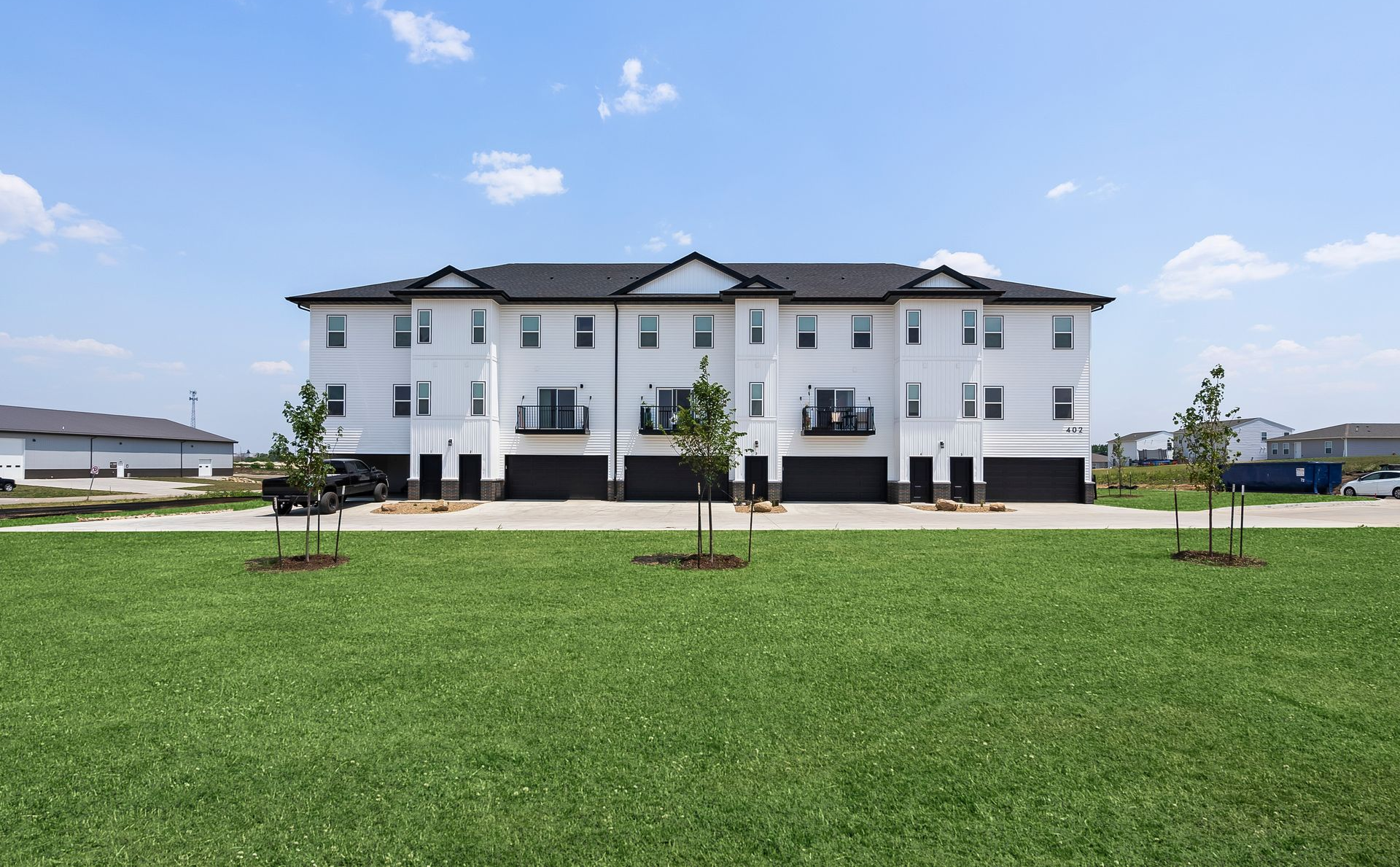 A three-story white townhouse building with dark roofs and garages sits behind a large green lawn under a sunny sky.