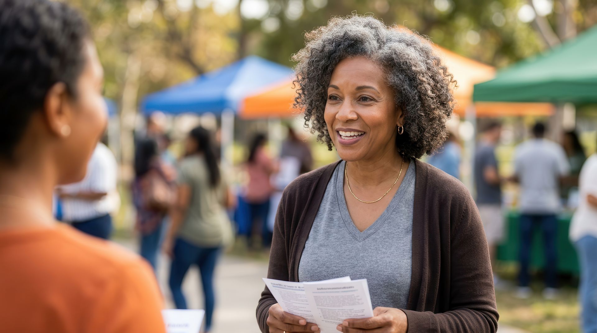 Woman holding papers and smiling at an outdoor event with colorful tents in the background