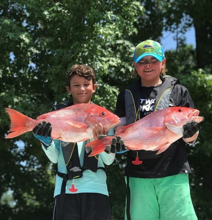 Two kids holding large red fish outdoors, wearing life vests and hats.