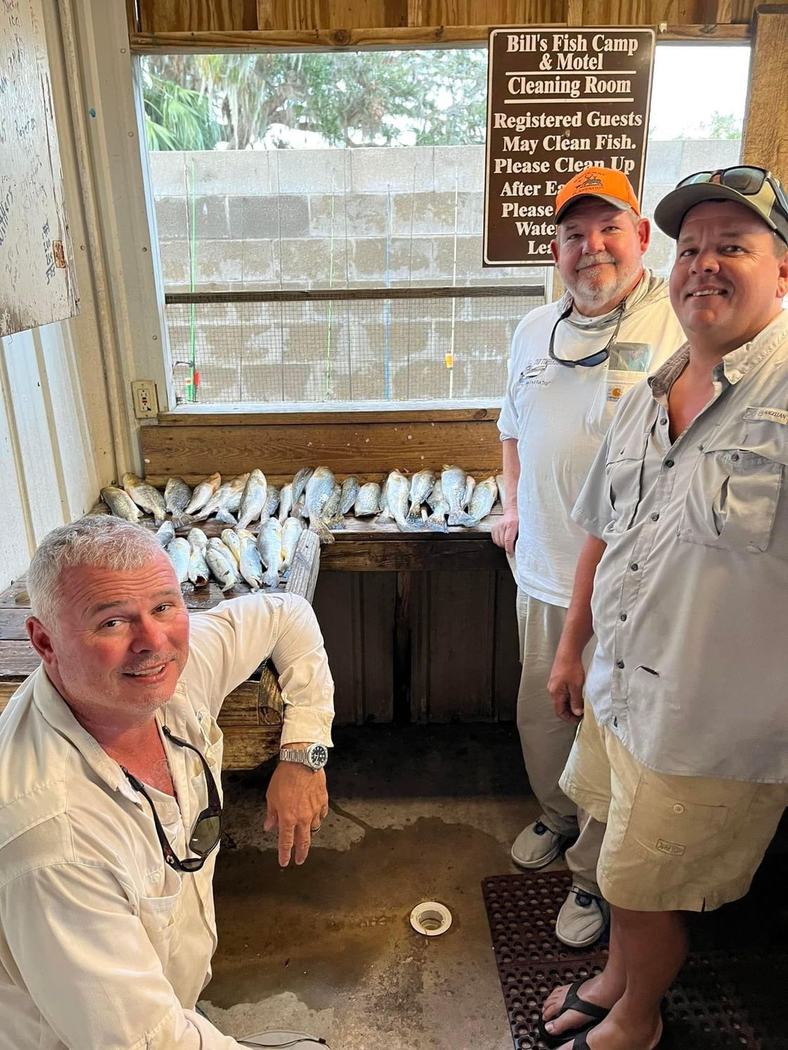 Three men in light shirts pose inside a rustic shed with a sign on the wall.