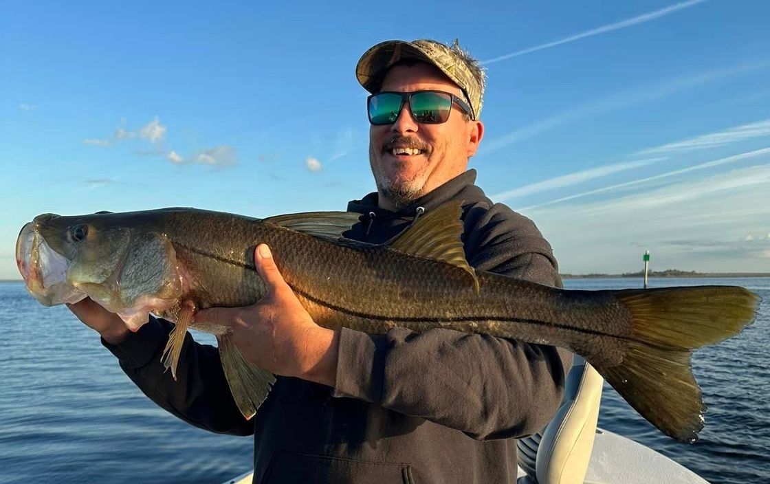 Man holding a large fish on a boat at sunset, with calm water and blue sky in the background