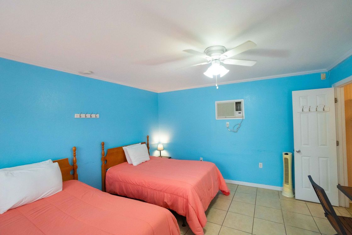 Bright blue bedroom with two coral beds, ceiling fan, wall AC, and tiled floor.