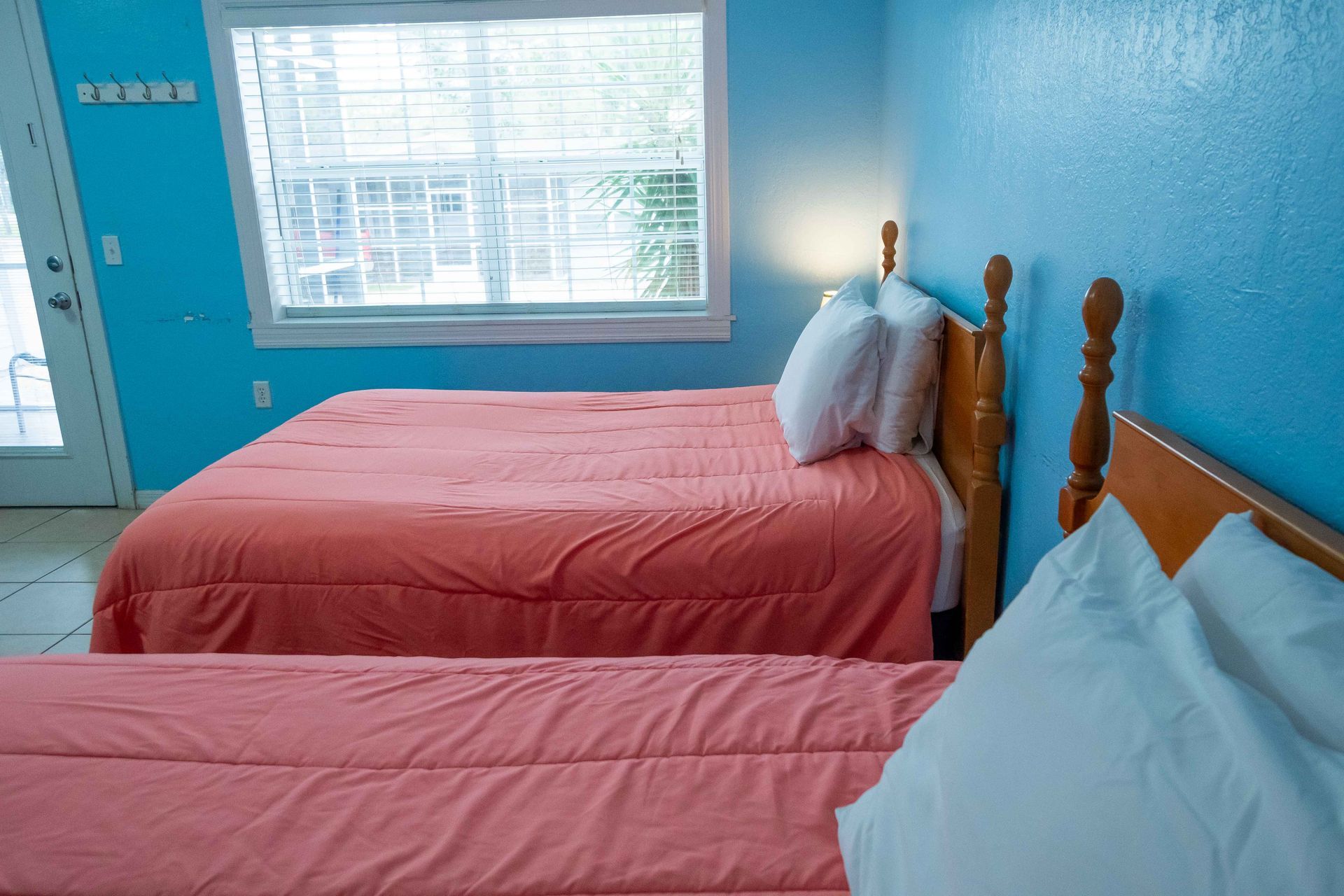 Bright blue bedroom with two twin beds, pink bedding, and a window letting in daylight