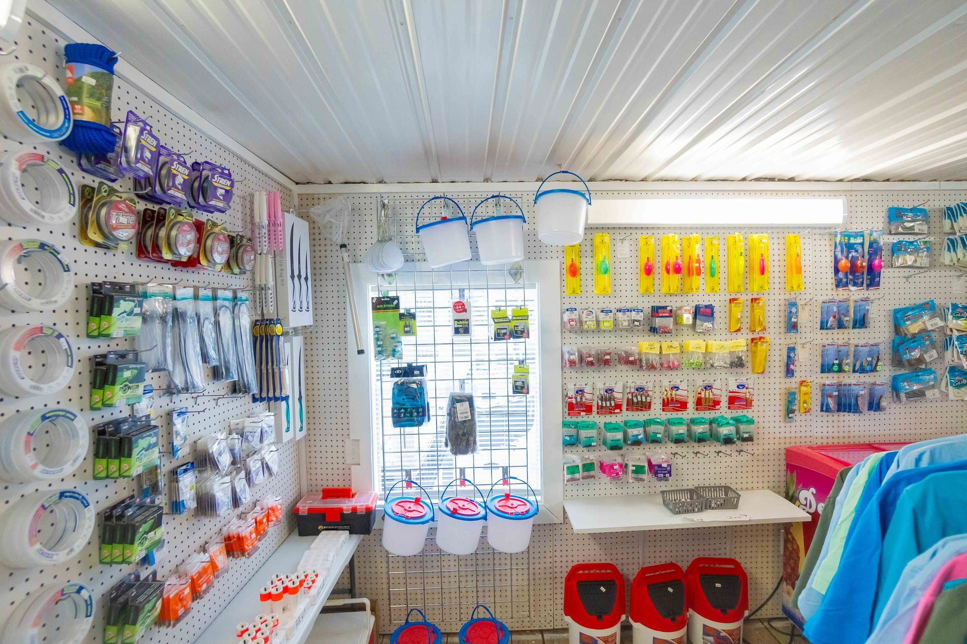 Bright store aisle with hanging pet supplies, colorful packaged items, and buckets near a windowed entrance