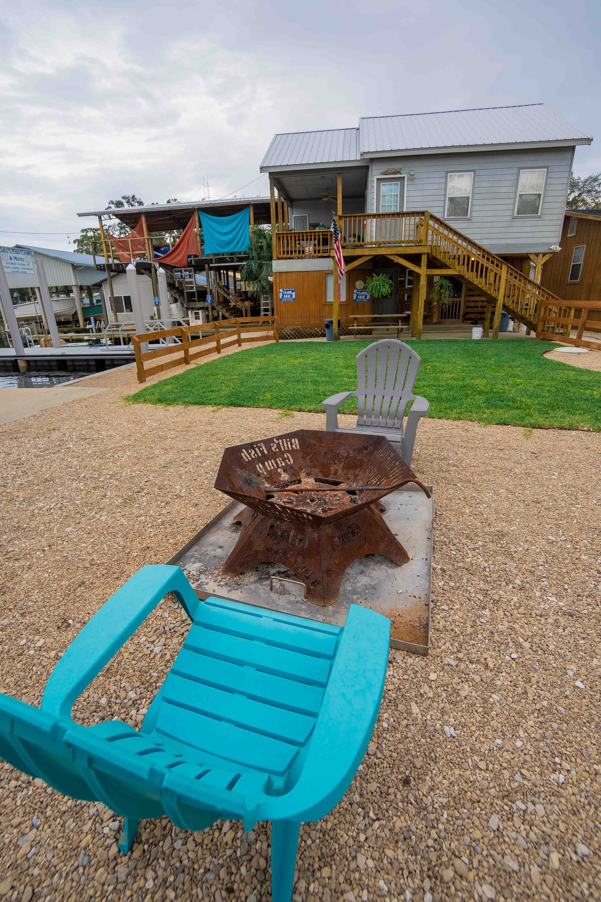 Outdoor seating area with turquoise chairs, a fire pit, gravel, grass, and a wooden deck overlooking water.