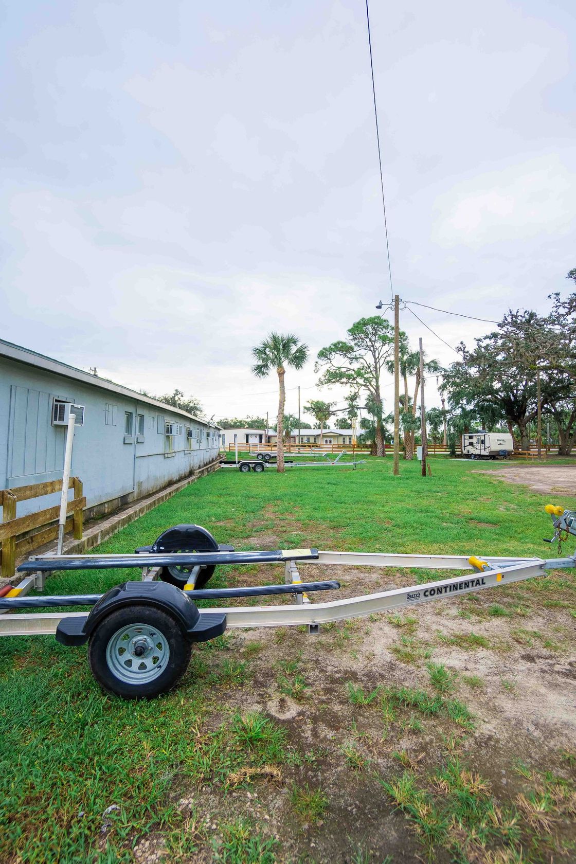 Small boat trailer on grass beside a mobile home with palm trees and a cloudy sky.