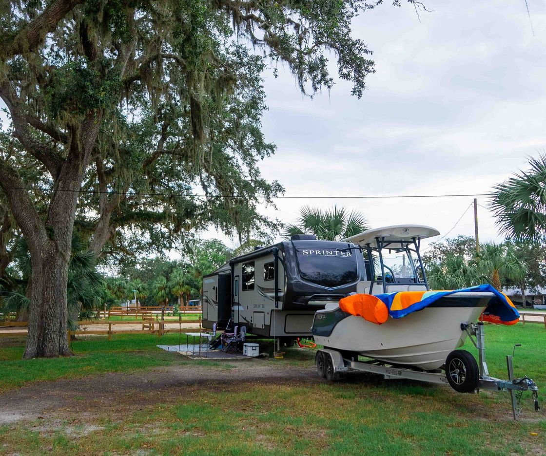 Camper trailer parked in a grassy campsite beside a small boat, under a large tree.