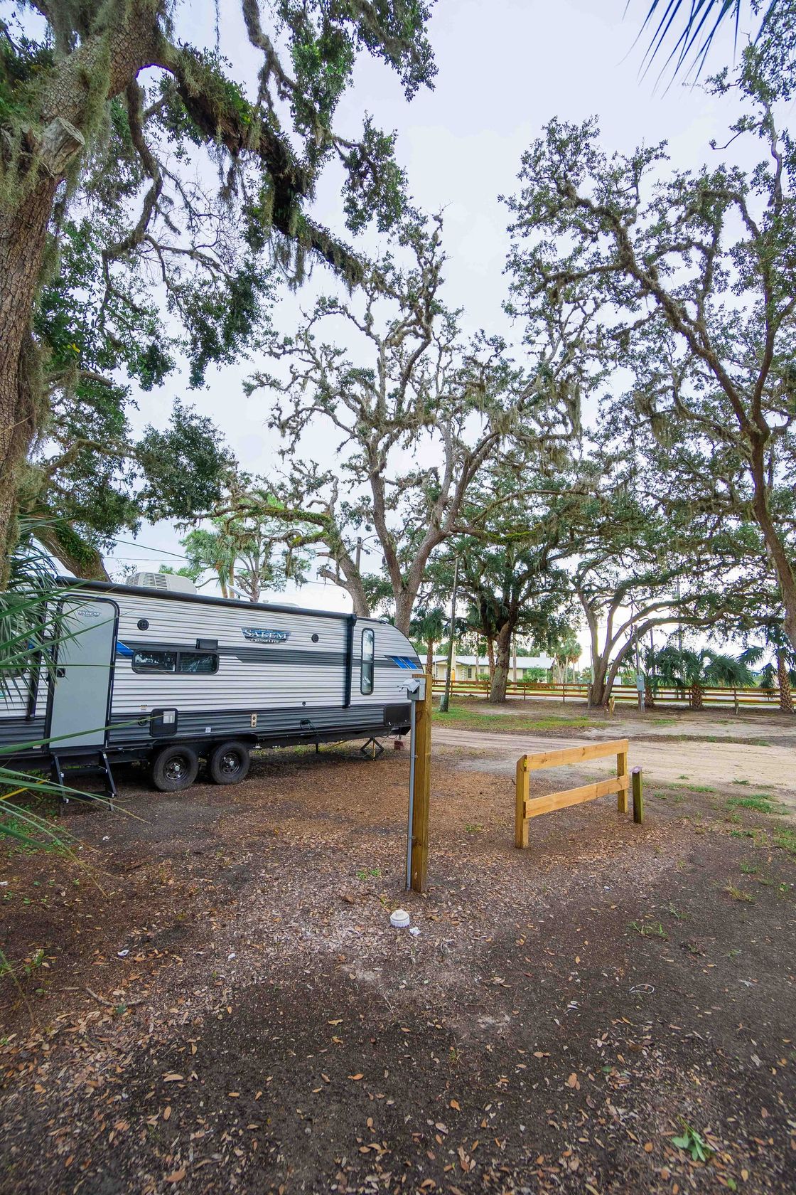 Camper trailer parked beside a dirt path under trees, with a yellow metal barrier in the foreground.