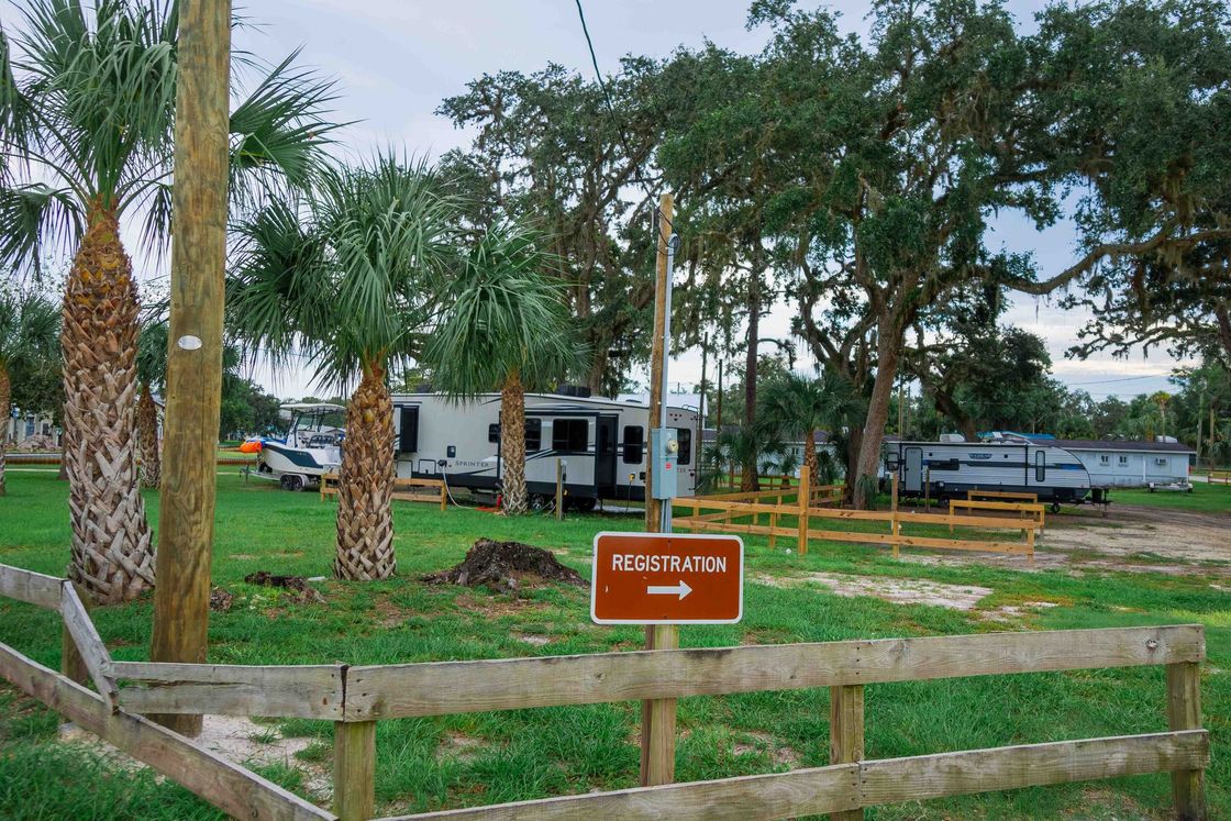 Sign reading “Admissions” by a grassy lot with trees, parked vehicles, and a fence.